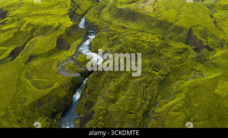 Aus der Vogelperspektive sehen Sie eine üppig grüne isländische Landschaft mit einem sich windenden Fluss, moosbedeckten Hügeln, zerklüfteten Klippen und weißen Stromschnellen, die sich durch Volks ziehen Stockfoto