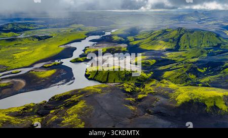 Aus der Vogelperspektive auf Islands vulkanisches Gelände mit einem sich windenden Fluss, grünen moosbedeckten Hügeln, schwarzem Sand und zerklüfteten Formationen unter einem Over Stockfoto