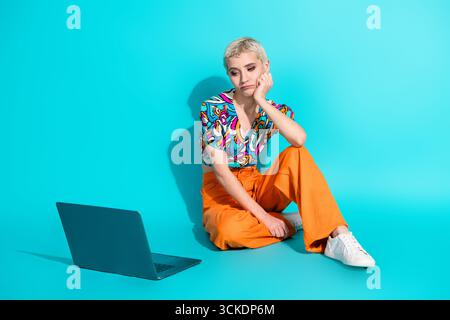 Eine stilvolle junge Frau sitzt in legerer Mode mit einem Laptop auf dem Boden vor leuchtendem blauem Hintergrund. Stockfoto