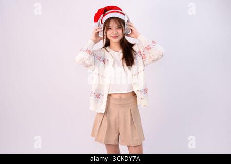 Lächelnde schöne Frau feiert weihnachten mit weihnachtsmütze und Kopfhörern in weißem Studiohintergrund Stockfoto