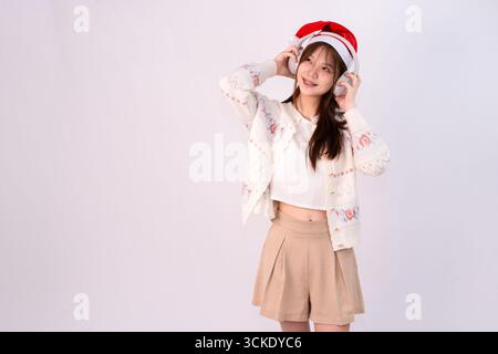 Lächelnde schöne Frau feiert weihnachten mit weihnachtsmütze und Kopfhörern in weißem Studiohintergrund Stockfoto