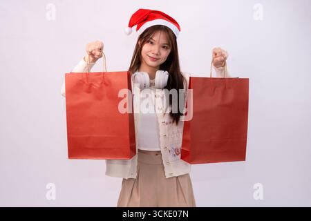 Junge schöne Frau im weihnachtsmann-Hut und Kopfhörer trägt rote Einkaufstaschen für weihnachtsfeiertage im weißen Studio-Hintergrund Stockfoto