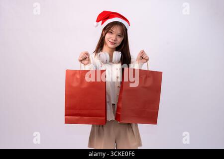 Junge schöne Frau im weihnachtsmann-Hut und Kopfhörer trägt rote Einkaufstaschen für weihnachtsfeiertage im weißen Studio-Hintergrund Stockfoto
