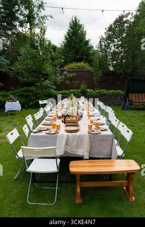 Gartenparty im Freien mit langem Tisch, Stühlen und Holzbank in üppigem Grün. Stockfoto
