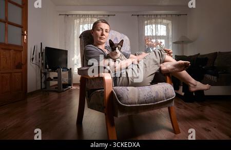 Eine Frau mittleren Alters mit grauen kurzen Haaren hält ihre kanadische Sphynx-Katze in den Armen und lächelt sanft in einem gut beleuchteten Wohnzimmer mit Holzböden und so Stockfoto