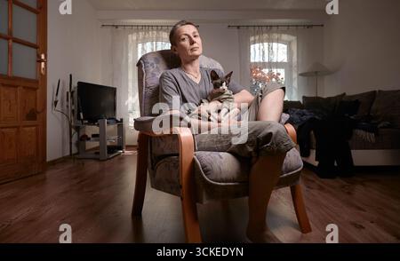 Eine Frau mittleren Alters mit grauen, kurzen Haaren sitzt bequem mit ihrer kanadischen Sphynx-Katze auf ihrem Schoß in einem modernen Wohnzimmer. Sie verkörpert Komfort, Frieden, A Stockfoto