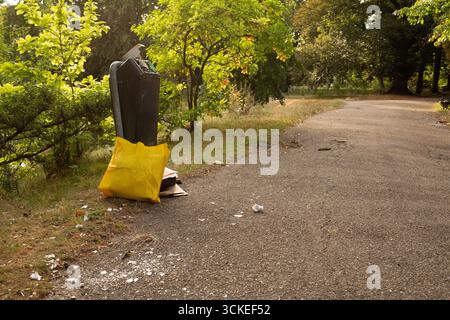 Mülltonne mit überlaufendem gelbem Beutel und verstreuten Müll auf Asphaltstraße im Park. Stockfoto