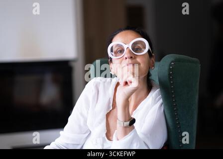 Selbstbewusste reife Frau mit schrulliger übergroßer Brille, die in einem grünen Sessel sitzt, ein weißes Hemd trägt, Hand am Kinn und in einem gemütlichen Zuhause nachdenklich aussieht. Stockfoto