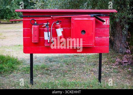 Hellrote Brandschutzstation für den Außenbereich mit mehreren Feuerlöschern, einem Schlauch, einer Schaufel, einem Haken und einem Metallschrank, der auf einem Holzbrett in einem Park montiert ist Stockfoto