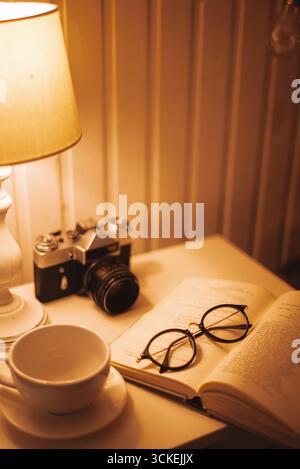 Ein gemütlicher Nachttisch mit offenem Buch, Lesebrille, einer alten Kamera und einer Tasse, alles in warmen Bädern Stockfoto