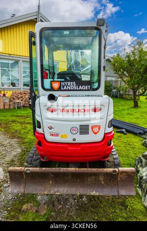 Kompakter Bagger Takeuchi TB225 mit Frontmesser, der auf grünem Rasen in der Nähe eines gelben Holzhauses mit Garten geparkt ist. Schweden. Stockholm. Stockfoto