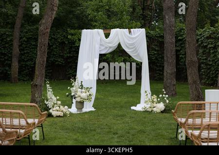 Ein wunderschöner Hochzeitsaltar befindet sich in einem ruhigen Garten, umgeben von Grün und mit weißen Vorhängen und Blumen dekoriert. Stockfoto