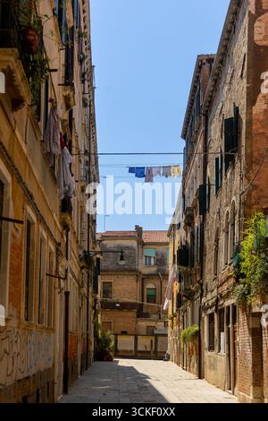 Wäschetrocknung über der schmalen Straße in Venedig. Farbenfrohe Kleidung hängt an den Linien zwischen rustikalen venezianischen Gebäuden in einer ruhigen Steinstraße und fängt sie ein Stockfoto