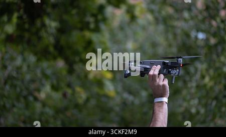 Nahaufnahme einer Person, die vor dem Flug eine Drohne in der Hand hält, mit verschwommenen grünen Bäumen im Hintergrund. Das Bild kann in Themen verwendet werden, die sich auf den Modus beziehen Stockfoto