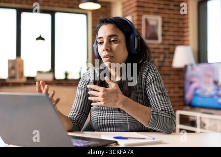 Indische Freiberuflerin sitzt drinnen mit Kopfhörern und spricht während eines Videogesprächs mit Kunden. Professionelle Frau arbeitet fernab von einer modernen Ziegelmauerwohnung mit Laptop und führt Online-Gespräche Stockfoto