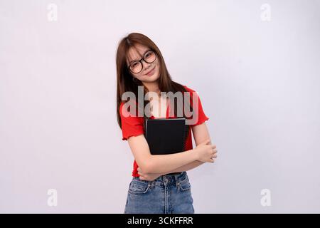 Junge Frau, die eine Brille trägt und ein Notebook hält, lächelnd in lässigem Outfit vor einem schlichten Hintergrund Stockfoto