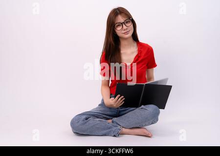 Eine fröhliche junge Frau mit Brille und lässiger Kleidung sitzt im Querbein, während sie im Studio liest Stockfoto