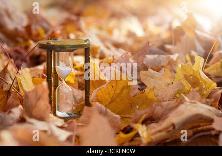 Alte Messing-Sanduhr steht draußen, umgeben von bunten Herbstblättern mit hellem Sonnenschein. Symbol der Zeit, Veränderung, Jahreszeiten, Nostalgie, Stockfoto