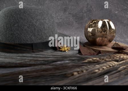 Moody Herbststill Life: Grauer Filzhut mit Blattgoldbrosche, Kerze in Kürbisform auf Holzbrett, weinrote Blätter und Weizenstiele. Stockfoto