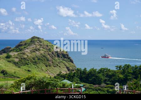 Eine rot-weiße Passagierfähre fährt an der vulkanischen Landzunge und dem blauen Horizont der Yongmeori-Küste vorbei. Stockfoto