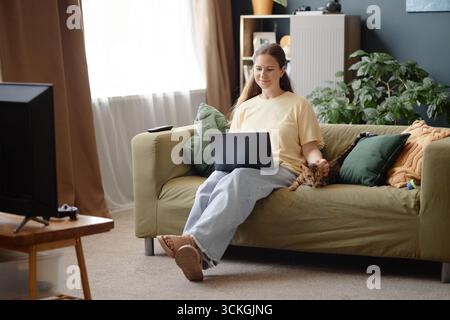 Junge Frau, die auf dem Sofa sitzt, mit Laptop und Katze im Haus streichelt Stockfoto