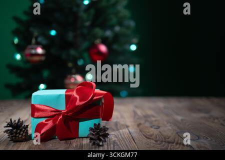 Ein wunderschön gewickeltes Geschenk sitzt auf einer Holzoberfläche, umgeben von Tannenzapfen, während ein dekorierter Weihnachtsbaum sanft im Hintergrund leuchtet. Perfekt für Urlaubsgrüße. Stockfoto