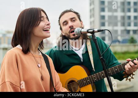 Kaukasische junge Frau, die neben dem kaukasischen jungen Mann in das Mikrofon singt und Akustikgitarre spielt und draußen singt, beide spielen gemeinsam Musik in urbaner Umgebung Stockfoto