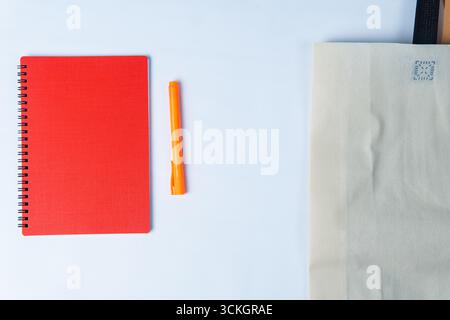 Minimalistisches Bild eines roten spiralförmigen Notizbuchs, eines orangefarbenen Stifts und einer beigefarbenen Stofftasche auf weißem Hintergrund – ideal für Themen wie Bildung, Büro, Kreativität, Stockfoto