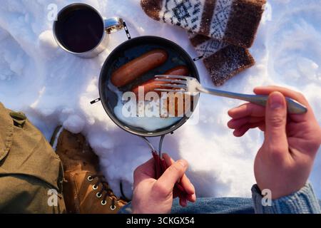 Kaukasischer junger Erwachsener, der eine Pfanne mit gekochten Würstchen und Spiegelei im Schnee hält, mit einer Gabel im Freien frühstückt, sichtbare Winterhandschuhe und Heißgetränk in der Nähe Stockfoto
