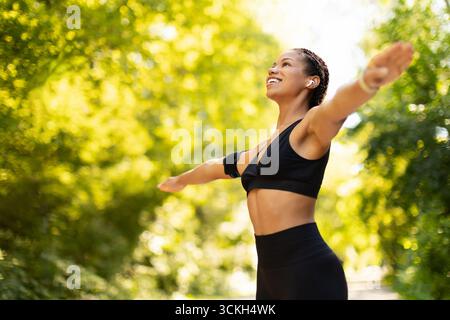 Fröhliche schwarze Frau mit weit geöffneten Armen in Sportbekleidung, die Outdoor-Workouts genießt Stockfoto