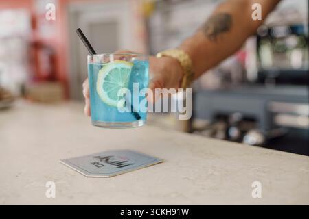 Crema, Italien - 25. August 2025 der Barkeeper legt erfrischenden blauen Cocktail mit Zitrone und Stroh auf die Bar neben der Achterbahn, damit Gäste ein drin genießen können Stockfoto