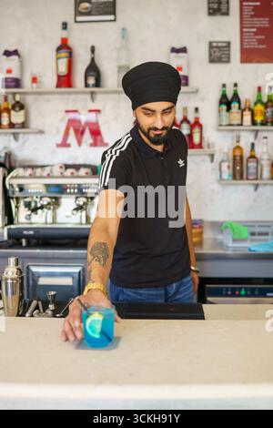 Crema, Italien - 25. August 2025 Ein junger Sikh Barista präsentiert dem Betrachter einen wunderschön garnierten blauen Cocktail, während er hinter einer Bar C arbeitet Stockfoto