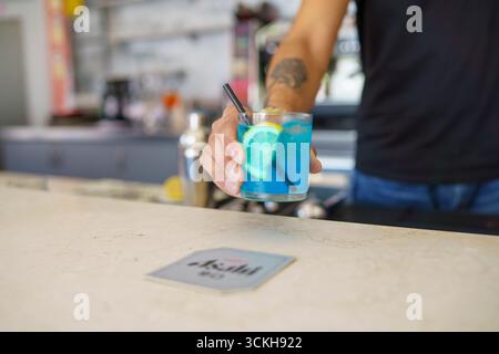 Crema, Italien - am 25. August 2025 bietet Bartender's Hand einen leuchtend blauen Cocktail, garniert mit Zitrone und Minze an einer sonnigen Bar Stockfoto