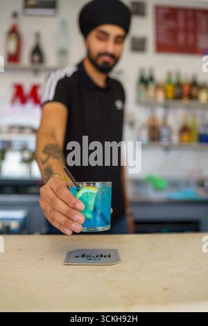 Crema, Italien - 25. August 2025 erfahrener Bartender mit Turban und Tattoos serviert erfrischenden Blauen Cocktail mit Zitronengeschmack und Eis in einer lebhaften Bar Stockfoto