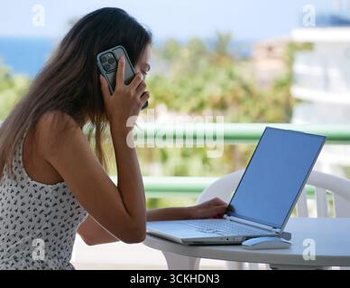 Freiberufliche Frau, die einen Laptop und ein Smartphone benutzt, während sie von einem Balkon mit tropischem Meerblick aus ferngesteuert arbeitet. Konzept des Multitasking, digitale Freiheit Stockfoto
