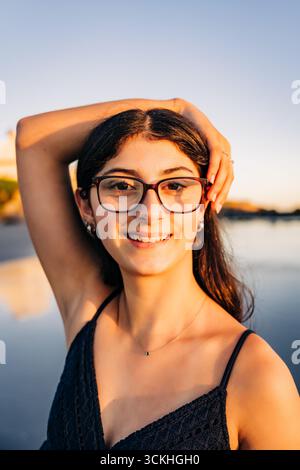 Lächelnde junge Frau mit Brille, die bei Sonnenuntergang am Strand posiert Stockfoto