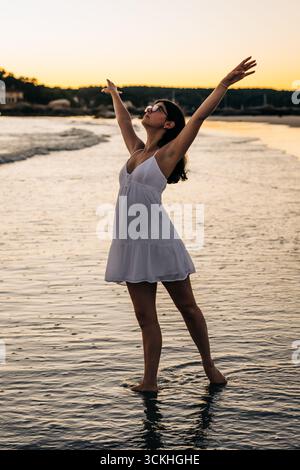 Junge Frau in weißem Kleid, die den Sonnenuntergang am Strand genießt Stockfoto
