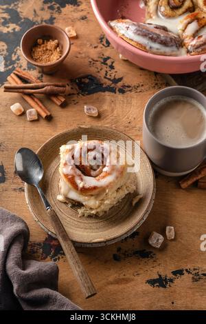 Frisch gebackenes zimtbrötchen, serviert auf Keramikplatte und Kaffeetasse auf strukturiertem Holzhintergrund. Vertikal Stockfoto