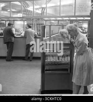 Verkaufsautomat 1957. Eine blonde Frau lehnt sich an den Automaten, der Schokoladentafeln verschiedener Art anbietet. Der typische, 1957 für Frauen beliebte Rock war ein breiter, ausgestellter A-Linie oder Kreisrock, der oft mit einem Gürtel an der Taille verschlungen wurde, um eine Sanduhr-Silhouette zu erzeugen. Es besteht aus Stoffen wie Baumwolle, Tüll oder Chiffon, fällt normalerweise auf mittlere Wadenlänge und wurde mit Unterröcken getragen, um mehr Volumen zu erhalten. Der Stil war lebendig, feminin und ikonisch mit der Mode des „New Look“ der 1950er Jahre. Conard Ref. 3496 Stockfoto