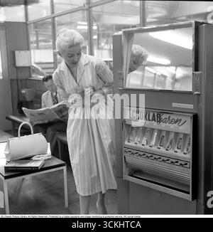 Verkaufsautomat 1957. Eine blonde Frau lehnt sich an den Automaten, der Schokoladentafeln verschiedener Art anbietet. Der typische, 1957 für Frauen beliebte Rock war ein breiter, ausgestellter A-Linie oder Kreisrock, der oft mit einem Gürtel an der Taille verschlungen wurde, um eine Sanduhr-Silhouette zu erzeugen. Es besteht aus Stoffen wie Baumwolle, Tüll oder Chiffon, fällt normalerweise auf mittlere Wadenlänge und wurde mit Unterröcken getragen, um mehr Volumen zu erhalten. Der Stil war lebendig, feminin und ikonisch mit der Mode des „New Look“ der 1950er Jahre. Conard Ref. 3496 Stockfoto