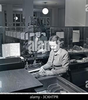 Das Innere einer Bank mit einer jungen Frau hinter dem Tresen, die den Kunden hilft. Man sieht sie beim Zählen von Banknoten mit einem Kunden am Kassenschalter. 1950. Kristoffersson Ref. AU71-6 Stockfoto
