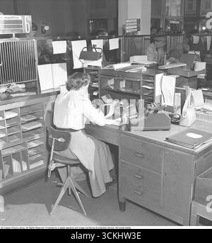 Das Innere einer Bank mit einer jungen Frau hinter dem Tresen im Bankbüro. Sie wird gesehen, wie sie einem Kunden hilft, der an der Kasse steht und sein Sparbuch hält. Ein Sparkassenbuch ist ein physisches Heft, das von Einzelpersonen verwendet wird, um Transaktionen auf einem Sparkonto zu erfassen, wie es in Banken und im Alltag vor dem digitalen Banking üblich ist. Es symbolisiert die Finanzgeschichte und die persönliche Sparkultur und ruft Nostalgie für traditionelle Bankpraktiken hervor. 1949. Kristoffersson Ref. AT21-1 Stockfoto