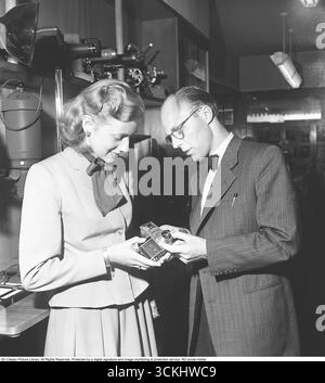 In einem Kamera- und Fotoausrüstungsladen wird 1949 einer jungen Frau die Kamera Exakta II gezeigt. Die 1949 vom Ihagee Kamerawerk in Dresden eingeführte Exakta II war eine kompakte 35 mm Spiegelreflexkamera, die auf dem Vorkriegsdesign von Kine Exakta aufbaut. Nach dem Zweiten Weltkrieg wurde es in begrenzten Mengen unter sowjetischer Kontrolle hergestellt und verfügte über einen linksseitigen Auslöser, einen Sucher auf Hüfthöhe und Kompatibilität mit Premium-Objektiven von Zeiss Jena und Meyer. In diesem Jahr ermöglichte ein Pentaprismuszubehör ein Betrachten auf Augenhöhe und erhöhte seine Attraktivität. Mit der Bezeichnung „Made in Germany“ wurde es zu einem begehrten Import in Stockfoto
