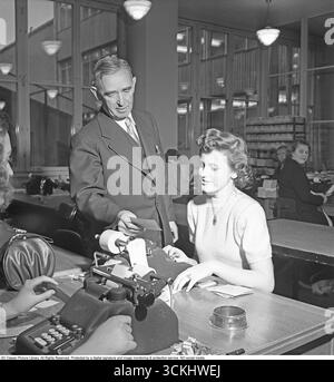 Ein Bankbüro in den 1940ern Weibliche Mitarbeiter arbeiten bei der Postspareinlage und erfassen Zahlungen, Abhebungen und Einzahlungen. Ein Mann steht neben einem Angestellten an einem mechanischen Taschenrechner und liest beide von einem kleinen Blatt Papier. Diese Szene spiegelt die wachsende Rolle der Frauen im Bankwesen und den Einsatz der frühen Bürotechnik in Europa der Nachkriegszeit wider. In den 1940er Jahren stieg die Zahl der Frauen, die in die Erwerbsbevölkerung einstiegen, einschließlich des Bankwesens, da nach dem Zweiten Weltkrieg die klerikalen Aufgaben erweitert wurden. Postspareinlagen, die in ganz Europa und darüber hinaus beliebt sind, boten zugängliche Finanzdienstleistungen an. (Kristoffersson, Ref. AU7 Stockfoto