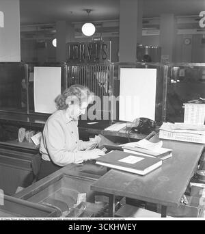 Das Innere einer Bank mit einer jungen Frau hinter dem Tresen im Bankbüro. Sie wird gesehen, wie sie einem Kunden hilft, der am Kassenschalter steht. Das Bild symbolisiert die Finanzgeschichte und die persönliche Sparkultur und ruft Nostalgie für traditionelle Bankpraktiken hervor. 1949 Kristoffersson Ref. AU72-7 Stockfoto