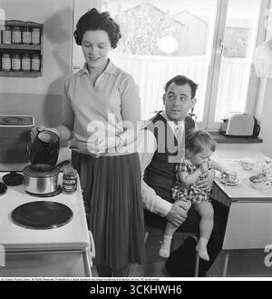 Sie bereitet den Kaffee 1961 zu. Ein Paar in der Küche, wo die Frau die neue Marke Nescafé benutzt, um leckeren Kaffee für ihren Mann zu kochen. Auf dem Küchentisch stehen Kaffeetassen mit dem von Sigvard Bernadotte entworfenen Virrvarr-Muster. Diese Szene spiegelt den Aufstieg von Instantkaffee und skandinavischem Design in den Haushalten der Nachkriegszeit wider. In den 1960er Jahren stieg der Instantkaffee weltweit an und Nescafé (1938 von Nestlé eingeführt) gewann wegen seiner Bequemlichkeit an Popularität, insbesondere in Europa der Nachkriegszeit, wo moderne Küchengeräte und -Produkte verwendet wurden. (Kristoffersson, Stockfoto