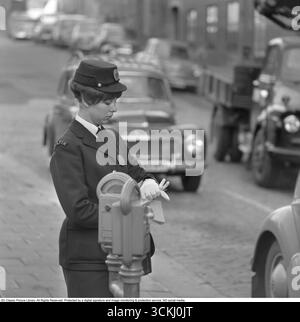 Es kostet Geld, falsch zu parken. Eine Frau arbeitet als Verkehrswächter und Parkwächter und hat die Aufgabe, Parkstrafen an einen Autofahrer zu verhängen, der nicht ordnungsgemäß geparkt hat oder die Parkzeit nicht bezahlt hat. Sie steht auf der Straße und schaut auf ihre Armbanduhr. Stockholm. 1963 Stockfoto