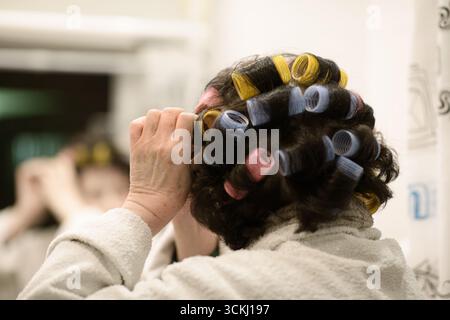 Eine ältere Frau in einem Bademantel legt helle Lockenwickler in ihr Haar, während sie vor einem Badezimmerspiegel steht, um einen ehrlichen Moment der persönlichen Pflege festzuhalten Stockfoto