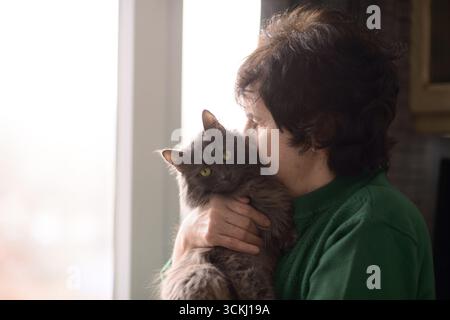 Eine ältere Frau mit grünem Pullover hält eine flauschige graue Katze in der Nähe eines Fensters. Das Bild fängt einen Moment der Wärme und der Gemeinschaft ein. Stockfoto