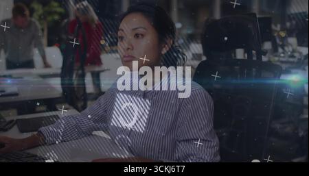 Asiatische Frau, die sich auf einen Desktop-Monitor im Großraumbüro konzentriert, mit digitalen Overlays Stockfoto